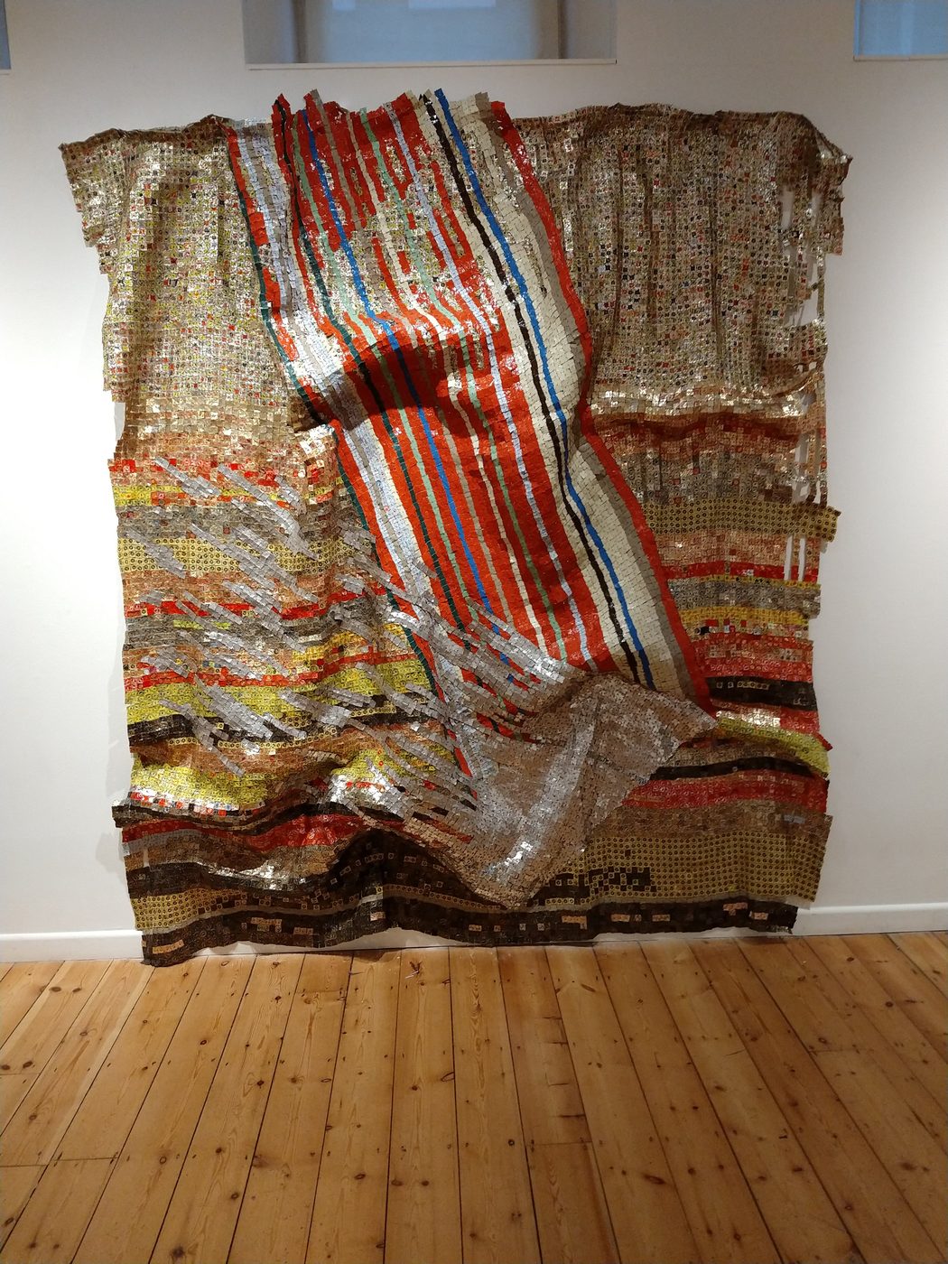 A large El Anatsui metal wall sculpture with red, white and blue stripes, draped across the gallery wall