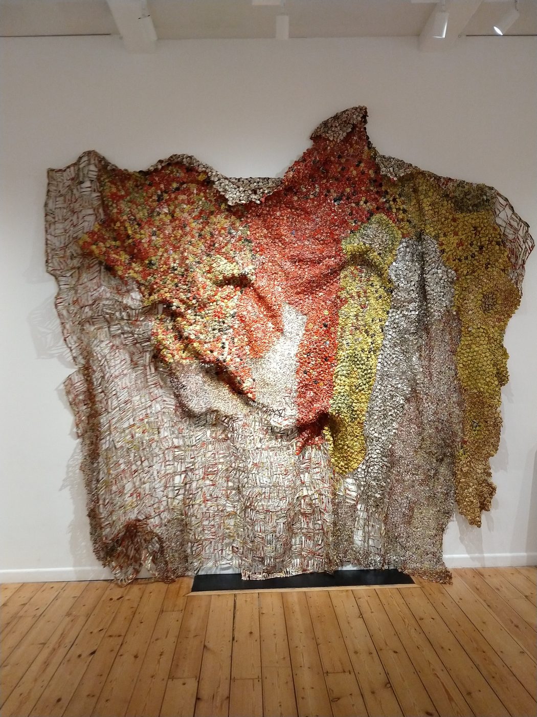 A wide El Anatsui metal wall sculpture in red, gold and silver, hung in folds